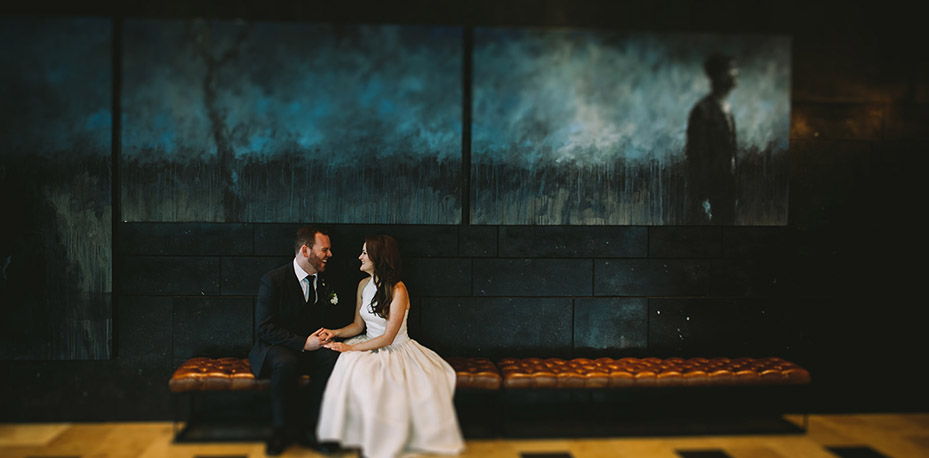 Ian and Rebecca sit and hold hands under hotel lobby artwork