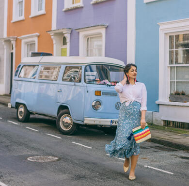 Woman walks down a street with a baby blue Volkwagen camper van in the background