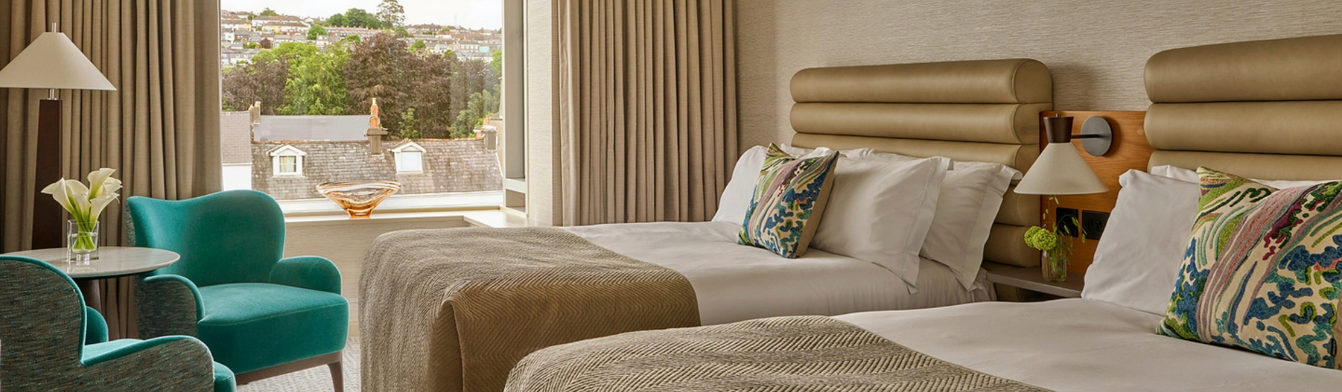 Classic Room at The River Lee Hotel featuring two double beds, a river view, and a seating area with two turquoise armchairs, a coffee table, and a lampshade.