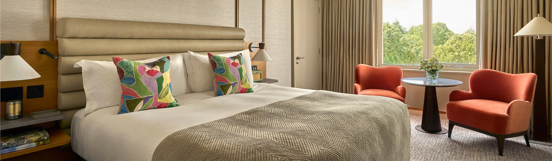 A bright Deluxe King Room at The River Lee hotel, featuring a large king-size bed and a seating area with two armchairs and a coffee table positioned in front of floor-to-ceiling windows.