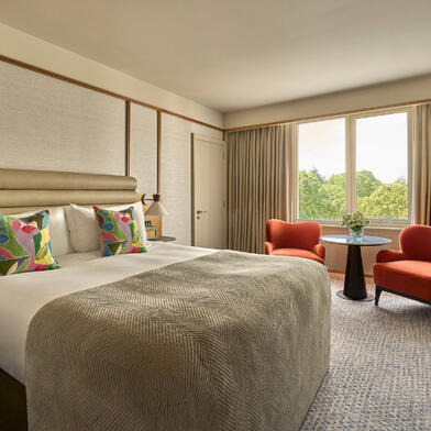 A bright Deluxe King Room showcasing a king bed styled with colorful decorative pillows, paired with two orange armchairs by the window.