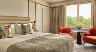 A bright Deluxe King Room showcasing a king bed styled with colorful decorative pillows, paired with two orange armchairs by the window.