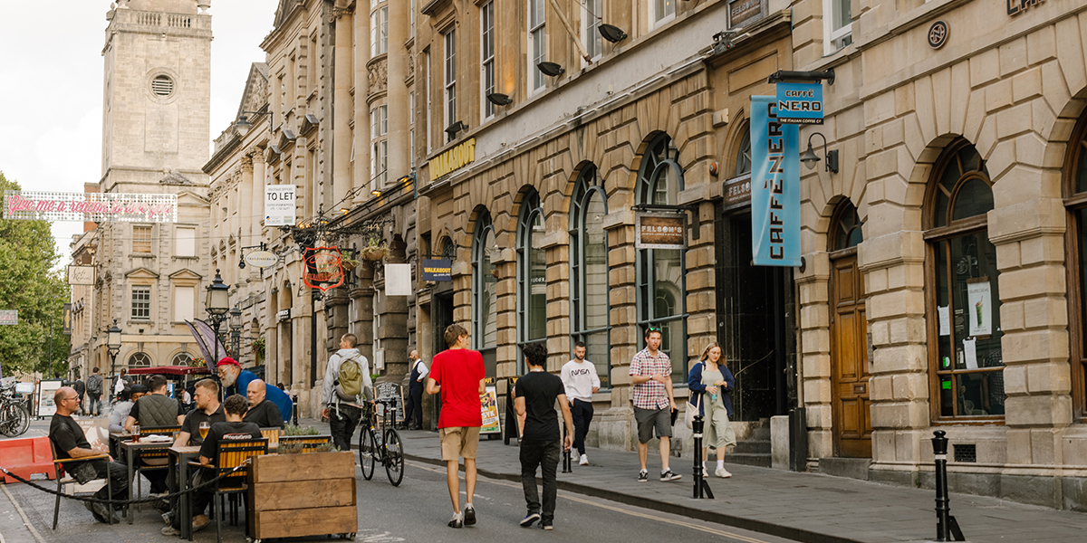 People dining at a lively outdoor restaurant terrace and pedestrians walking down a busy street in central Bristol.