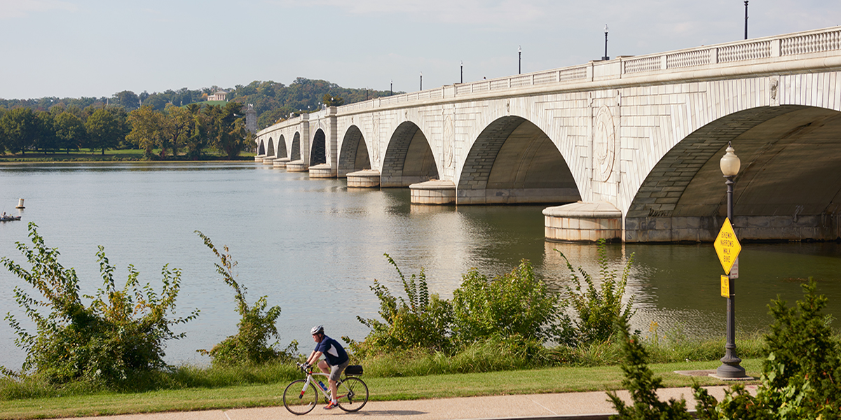 Washington, DC waterfront featuring a cyclist on a riverside bike path alongside the Potomac River and a bridge in the background.