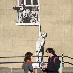Two tourists admiring a vibrant street art mural on a building in Bristol, depicting a man and a woman looking out from a painted window. The lively artwork adds to the city's creative atmosphere.
