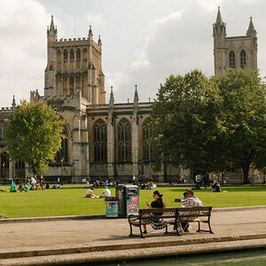 Two people relaxing on a park bench under the spring sunshine, with a grand, historic building visible across a well-maintained lawn, capturing the tranquility of a seasonal stroll.