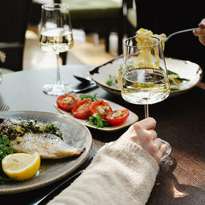 Close-up of a plate of perfectly cooked fish and a glass of wine at The River Grille, enjoyed by a female diner in the background