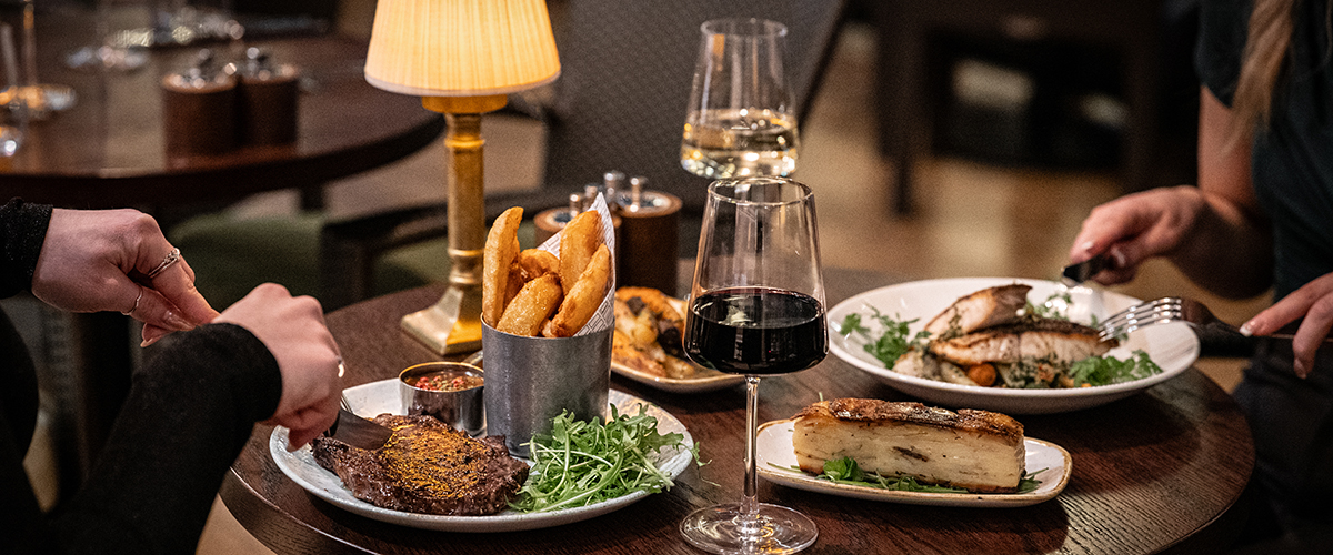 Two women dine at The River Grille, their hands visible on the table, which holds a steak, a fish dish, and glasses of wine under romantic, warm lighting.