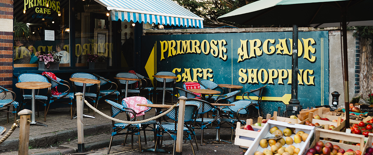 An autumnal scene in Clifton Village showing a farm shop with fresh vegetables and baskets outside, with the painted walls of Primrose Cafe on the left.