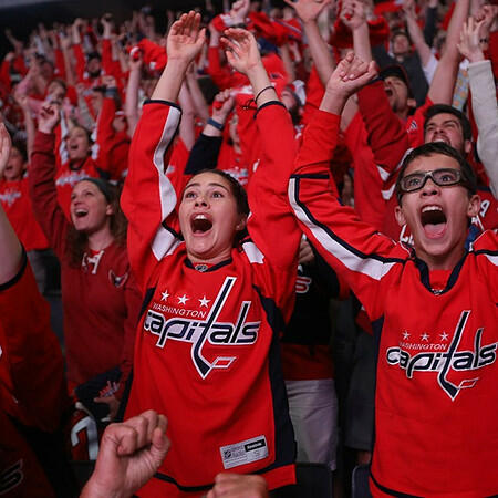 Vibrant scene at a Capitals hockey game with a sea of fans in red 'Rock the Red' gear celebrating a scoring play with arms raised and cheers.
