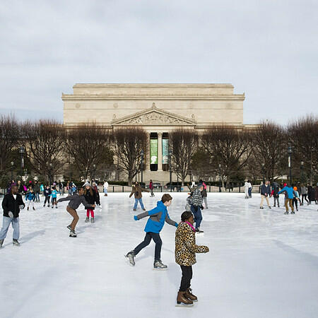 A lively winter scene at a DC ice rink featuring kids skating in the front and people of all ages gliding across the ice with views of the city in the background.