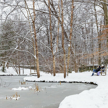 A snowy winter scene featuring two people on a bench facing a frozen lake