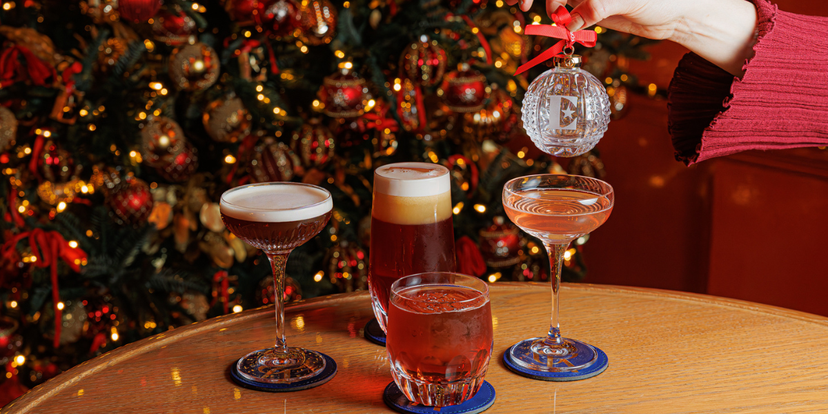 Detailed image showcasing The Doyle Collection's cocktails, served in four unique Waterford crystal glasses. The setting is festive, complete with a Christmas tree and a person holding a complementary crystal bauble.