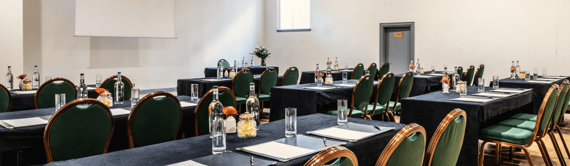 Meeting in The Bloomsbury Hotel's Tavistock room. The room is arranged with tables and green chairs in rows, with a black tablecloth on each table. Each table has three chairs and is spaced to allow people to pass through the rows. Bottles of water are on the tables.