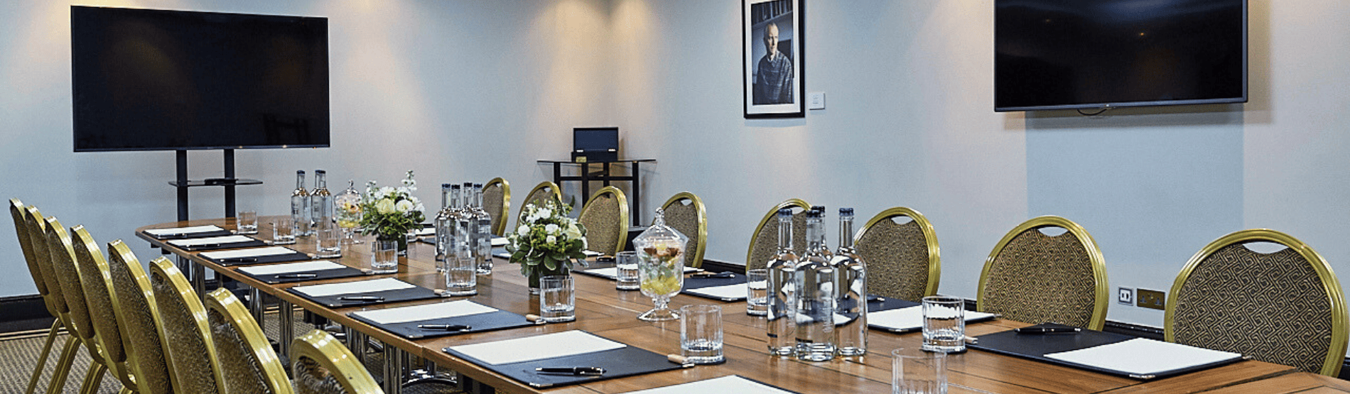 A corporate meeting room setup in The Bedford Studio at The Bloomsbury Hotel. The room features a long table with a vase of flowers, bottles of water, pens, and paper in front of each chair. There are two large TVs in the room for presentations.