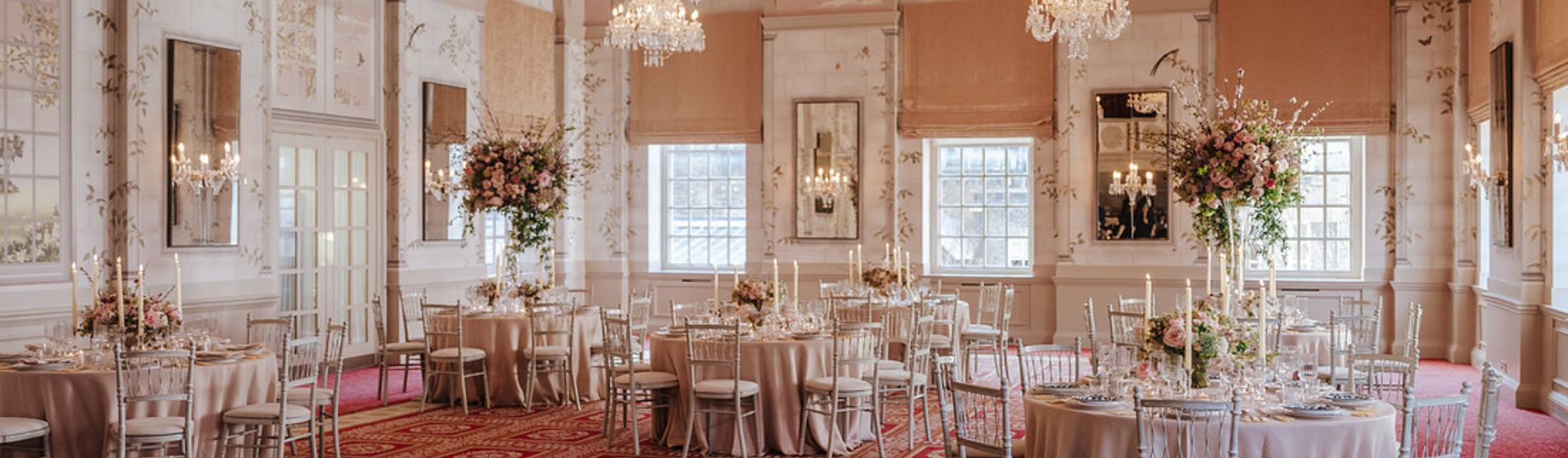 Queen Mary Hall prepared for a special occasion, with round dining tables styled with full place settings and tall vases of flowers. The room’s historic details and statement chandeliers add a sense of luxury and romance.