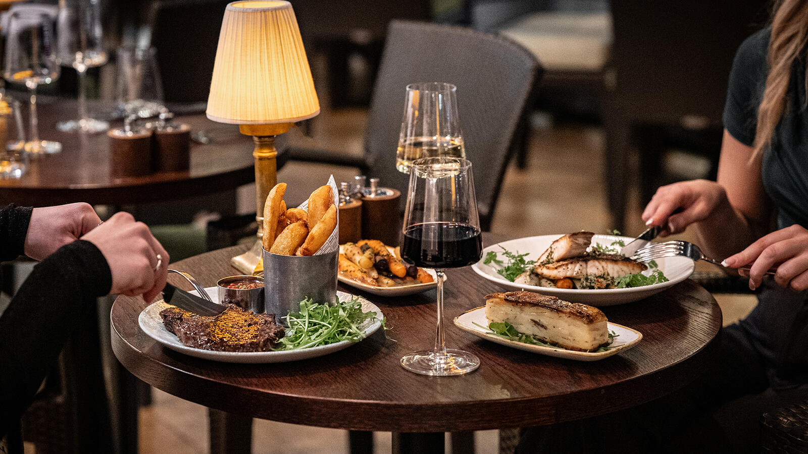 Elegant dining at Ricks Restaurant in The Bristol Hotel. Two guests savoring their steak and fish main courses, complete with wine pairings.