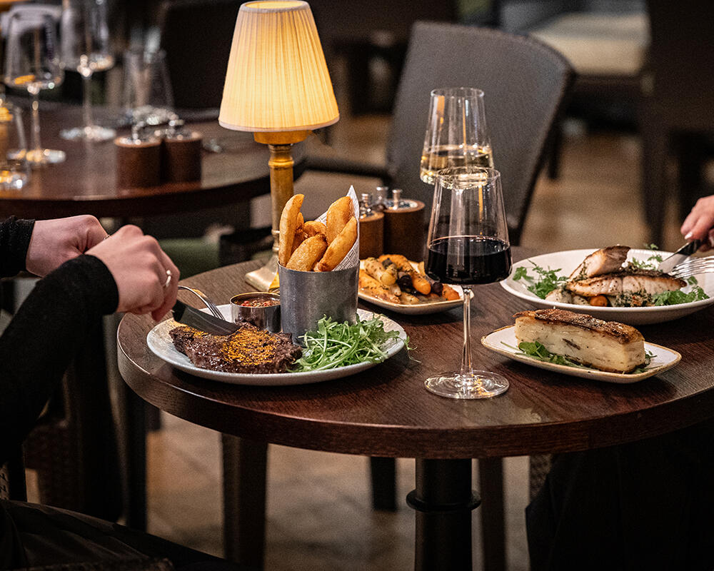 Elegant dining at Ricks Restaurant in The Bristol Hotel. Two guests savoring their steak and fish main courses, complete with wine pairings.
