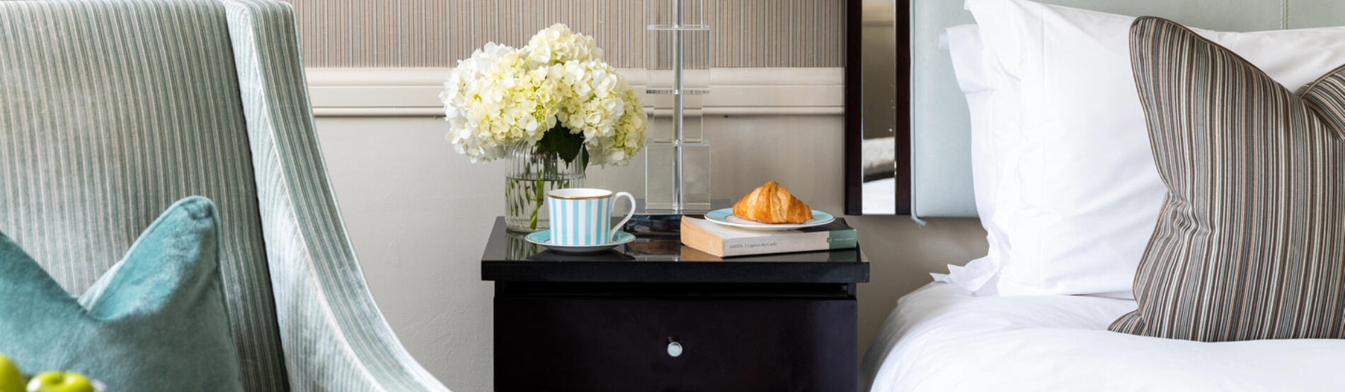 A cozy hotel room at The Bristol, ready for a winter escape. A neatly made bed is visible, with a side table holding a coffee, croissant, a book, and a white flower and a comfortable armchair with a turquoise pillow beside.