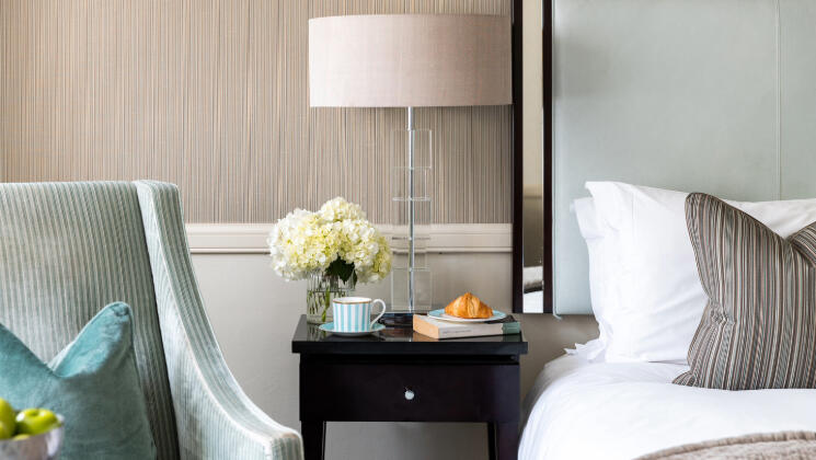 A cozy hotel room at The Bristol, ready for a winter escape. A neatly made bed is visible, with a side table holding a coffee, croissant, a book, and a white flower and a comfortable armchair with a turquoise pillow beside.