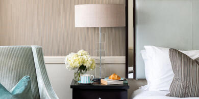 A cozy hotel room at The Bristol, ready for a winter escape. A neatly made bed is visible, with a side table holding a coffee, croissant, a book, and a white flower. A comfortable armchair with a turquoise pillow.