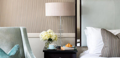 A cozy hotel room at The Bristol, ready for a winter escape. A neatly made bed is visible, with a side table holding a coffee, croissant, a book, and a white flower. A comfortable armchair with a turquoise pillow.