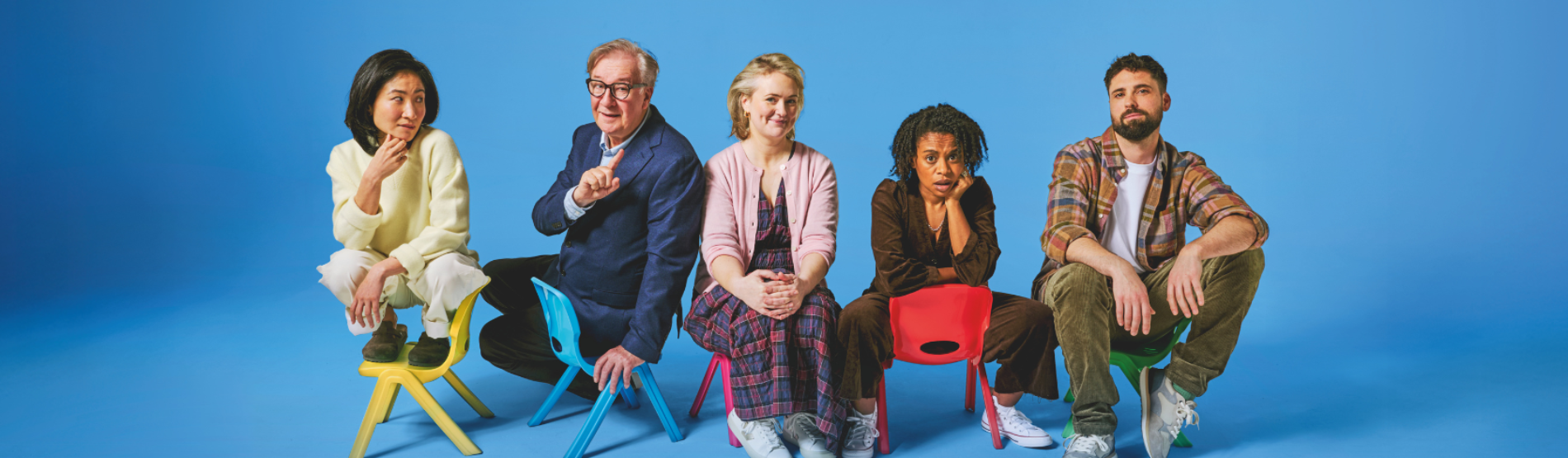 Cast members of the Gate Theatre’s production of Eureka Day sitting in small, colorful children's chairs against a vibrant blue background. The actors are posed in various expressions, capturing the play's comedic atmosphere.