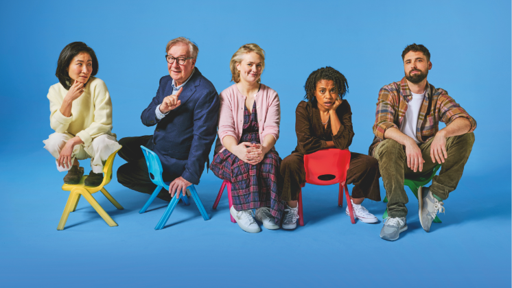 Cast members of the Gate Theatre’s production of Eureka Day sitting in small, colorful children's chairs against a vibrant blue background. The actors are posed in various expressions, capturing the play's comedic atmosphere.