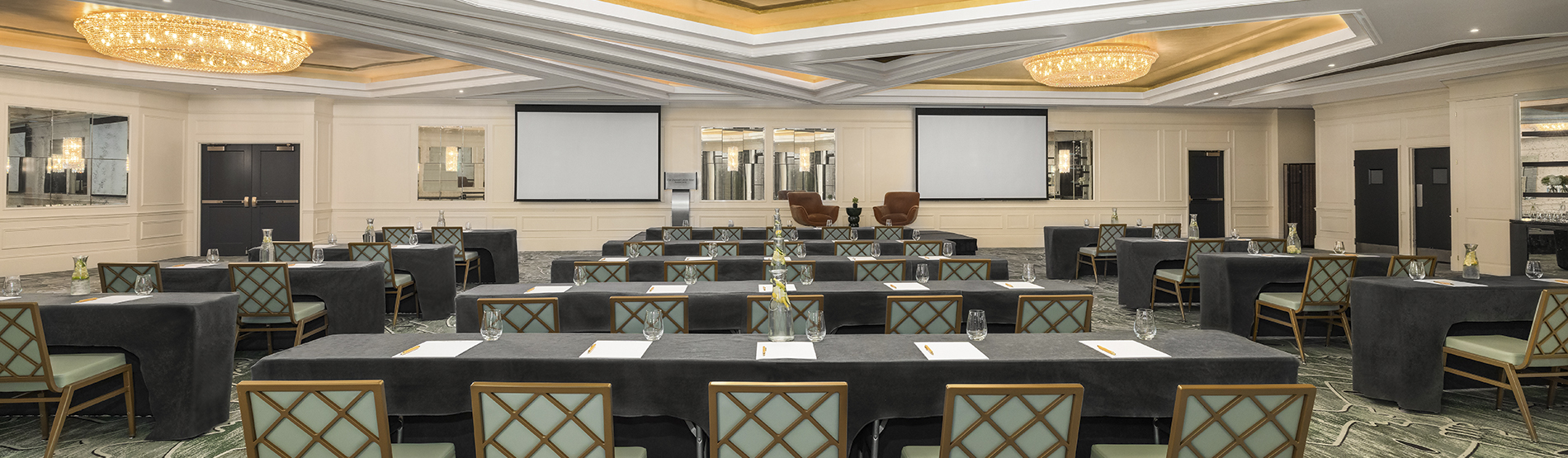 A wide view of The Dupont Ballroom set in a classroom-style arrangement. Orderly rows of long tables and chairs face the front of the hall, illuminated by the warm glow of the room’s signature circular ceiling lights.