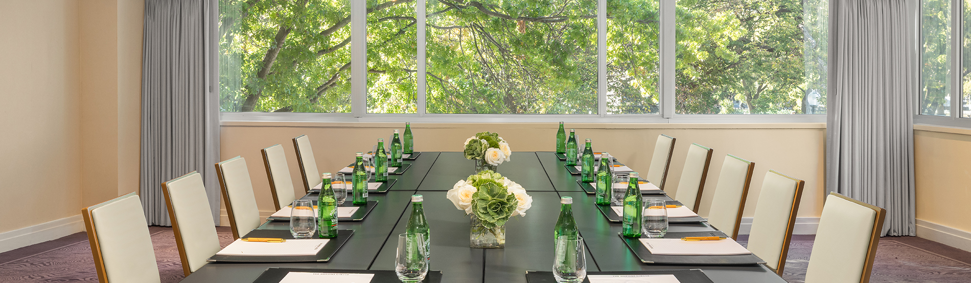 Georgetown Room conference setup at The Dupont Circle hotel featuring a boardroom table with water, notes, and white-and-green floral vases. Large windows in the background provide a scenic view of tree-lined streets in DC.