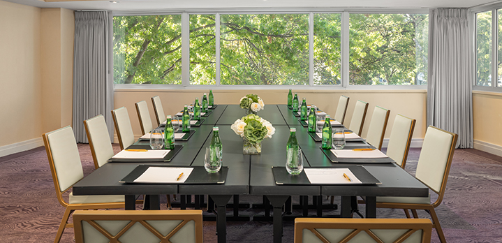 Georgetown Room conference setup at The Dupont Circle hotel featuring a boardroom table with water, notes, and white-and-green floral vases. Large windows in the background provide a scenic view of tree-lined streets in DC.