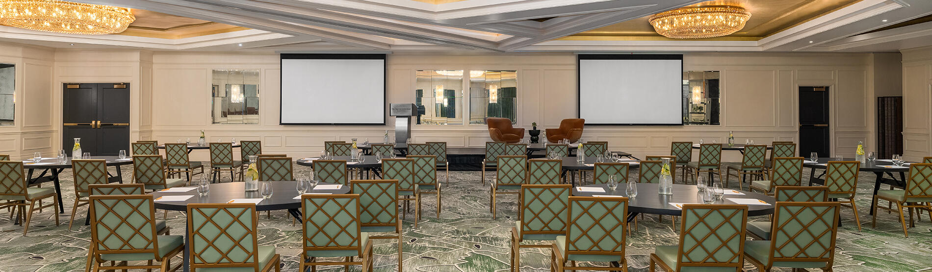 A wide shot of the Dupont Ballroom Crescent in The Dupont hotel arranged for a meeting. Round tables and chairs are neatly spaced throughout the room, illuminated by a large, ornate circular light fixture that serves as a central focal point.