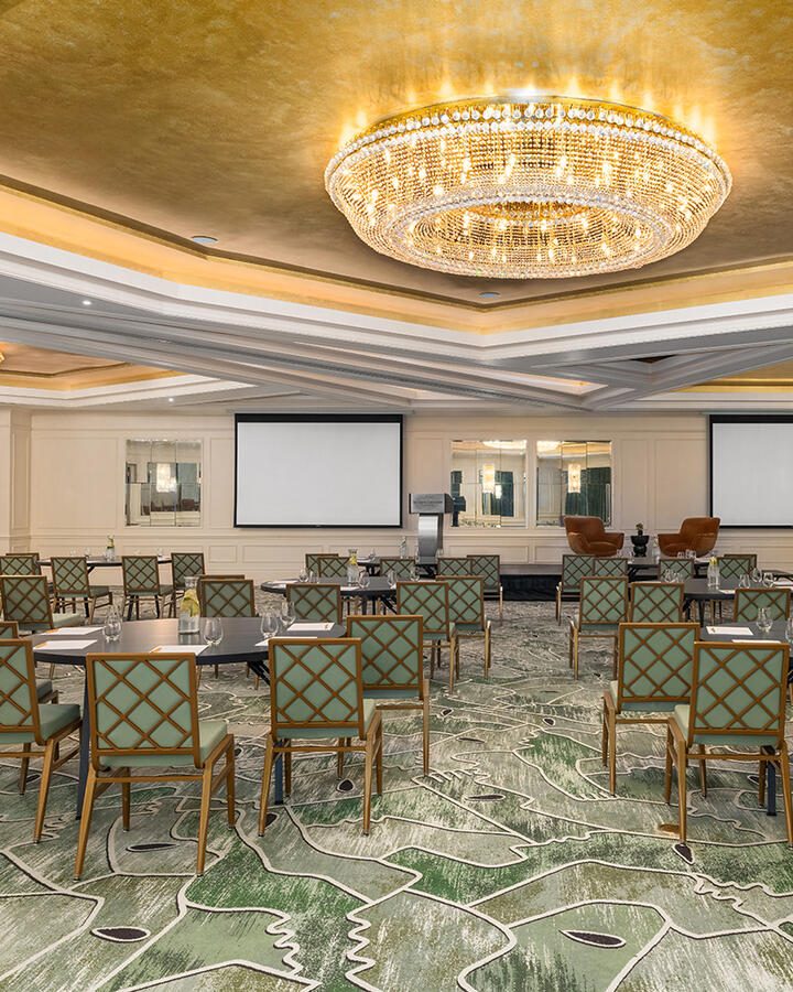 A wide shot of the Dupont Ballroom Crescent in The Dupont hotel arranged for a meeting. Round tables and chairs are neatly spaced throughout the room, illuminated by a large, ornate circular light fixture that serves as a central focal point.