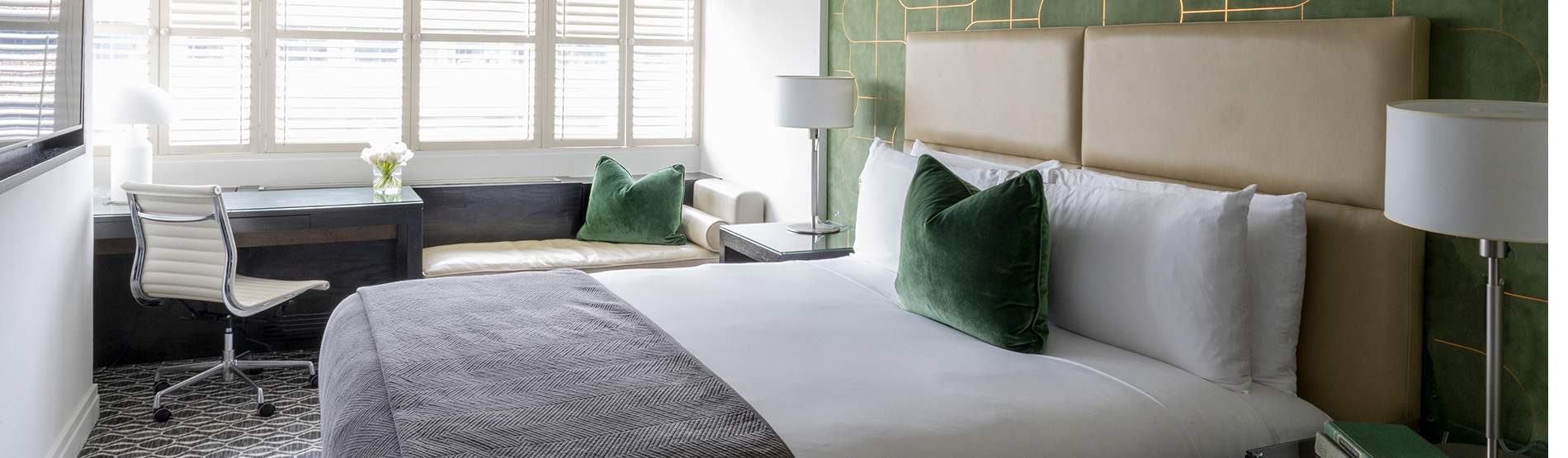 An accessible room at The Dupont Circle hotel. The room features a bed with white sheets and green pillow on the bed, a desk, and a side table, all with lamps. The walls are also decorated with a green accent color.