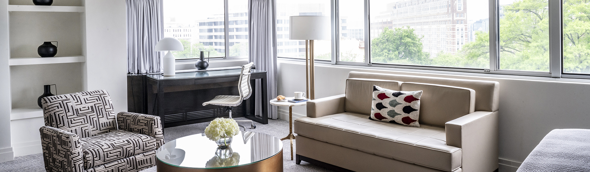 A bright and spacious pet-friendly suite furnished with a desk, a sofa, and an armchair at The Dupont Circle hotel. The room is filled with natural light from large windows that offer a view of Washington, D.C. 