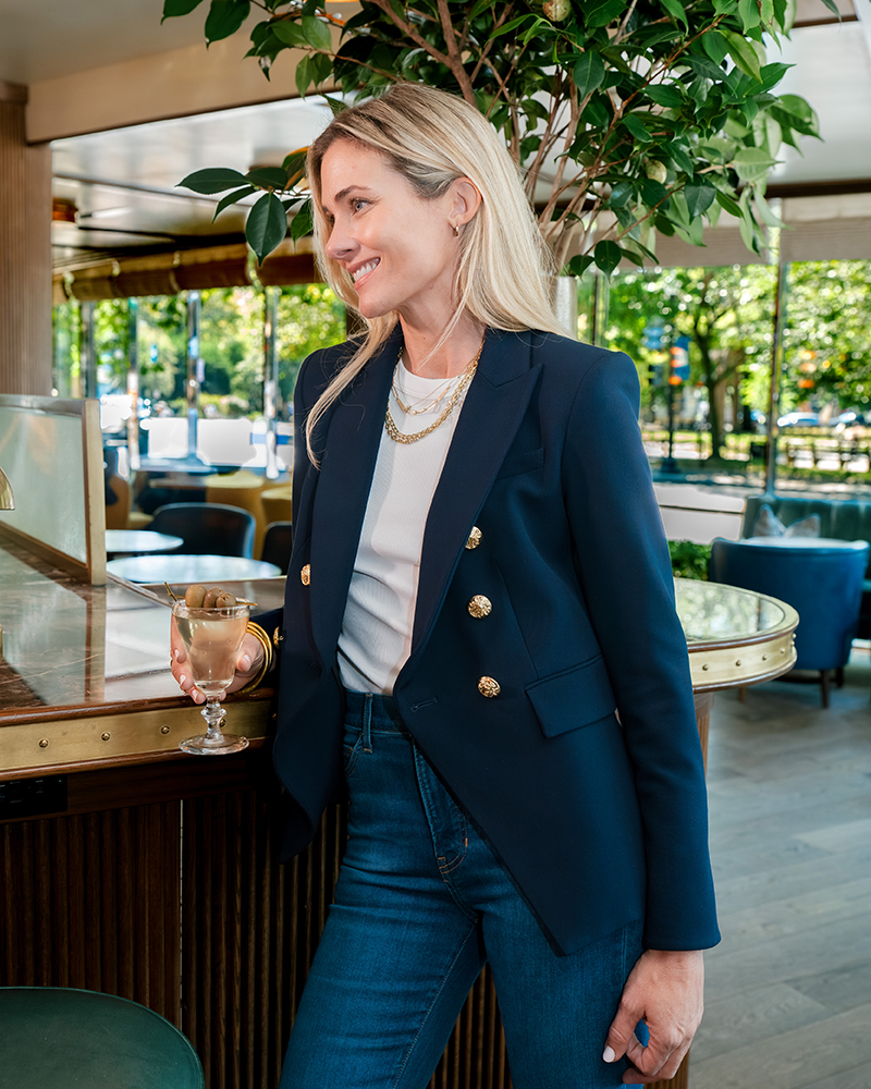 Veronica Beard, wearing a navy blazer, is shown inside the bar at The Dupont Circle Hotel, holding a glass.