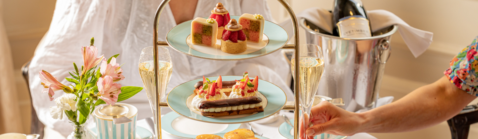 A bright, upscale afternoon tea service for two at The Kensington Hotel. Two ladies are seated at a beautifully set table, mid-conversation with glasses of champagne in hand, featuring a three-tier stand filled with pastries and sandwiches