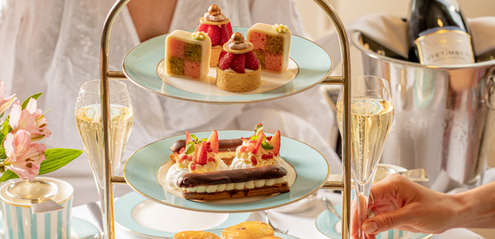 A bright, upscale afternoon tea service for two at The Kensington Hotel. Two ladies are seated at a beautifully set table, mid-conversation with glasses of champagne in hand, featuring a three-tier stand filled with pastries and sandwiches