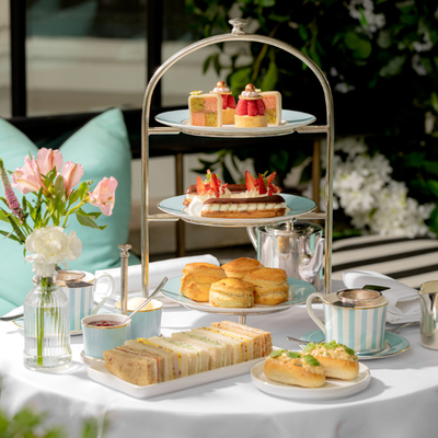 Luxury British afternoon tea experience inside the elegant dining room of The Kensington Hotel in London, showcasing a beautifully presented 3-tier stand of savory sandwiches, artisanal scones, and sweet pastries next to a silver tea set
