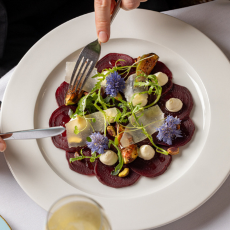  A person dining at The Kensington Hotel London is eating a beautifully plated dish of beetroot carpaccio garnished with figs, cheese shavings, cornflowers, and pistachios.