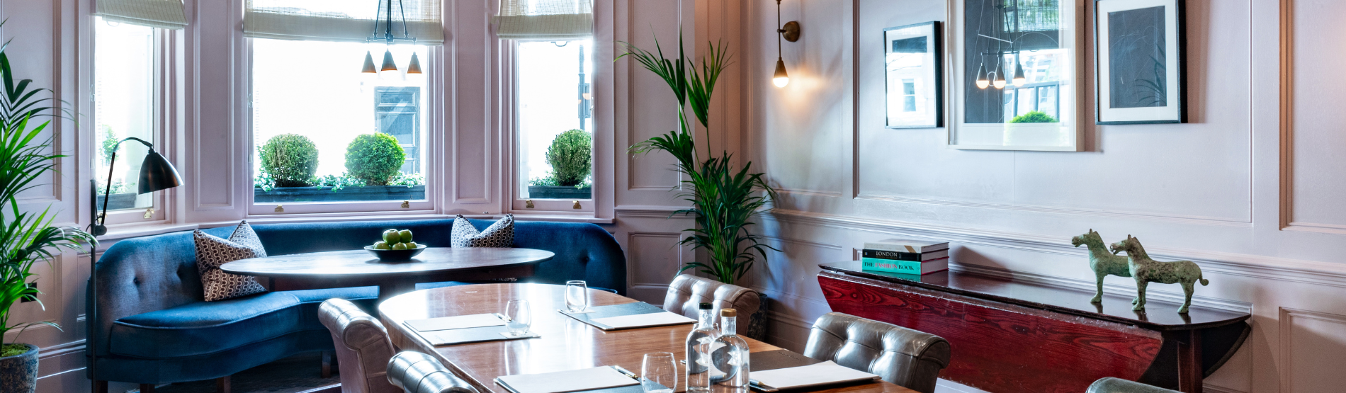 A bright and elegant room called The Study at Town House, The Kensington, featuring classic decor, modern furniture, a large wooden table set for a meeting or dining, blue velvet banquette seating by bay windows, potted plants, and tasteful artwork—ideal for private dining, meetings, and receptions.