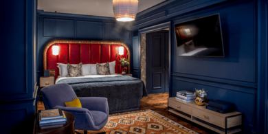 Luxury studio suite at The Marylebone Hotel. The room, decorated with rede and blue accents, features a plush bed, a stylish armchair, and a tv.