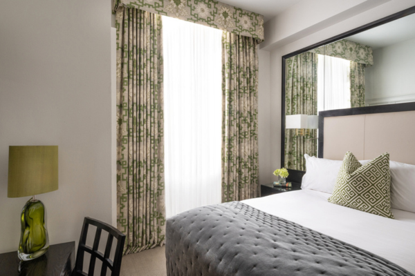 Classic Room 1 Queen Bed at The Kensington. The room is well-lit and features a bed with white bedding and green pillows, alongside complementary green draperies and a green desk lamp.