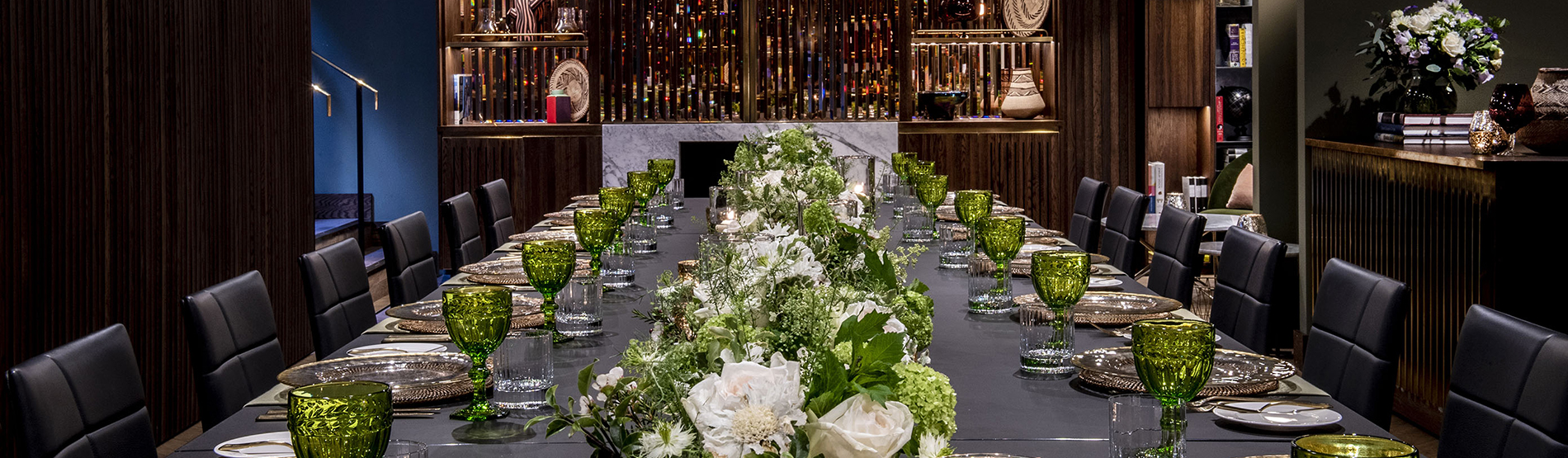 Sophisticated long dining table at The Marylebone set with polished plates, glassware, and cutlery, featuring elegant white and green floral arrangements under warm, moody lighting.
