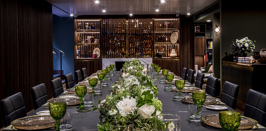 An elegantly set long dining table featuring polished glassware, plates, and cutlery arranged perfectly alongside vibrant white and green floral centerpieces under sophisticated, dimmed lighting.