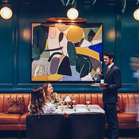 Two women enjoying pre-theatre dining in Cork at The River Club grill, as a waiter serves wine.