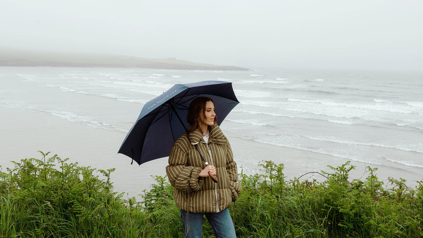 Louise O'Neill standing on the West Cork coast with an umbrella against a moody gray sky, showcasing the wild beauty of Cork's seaside.