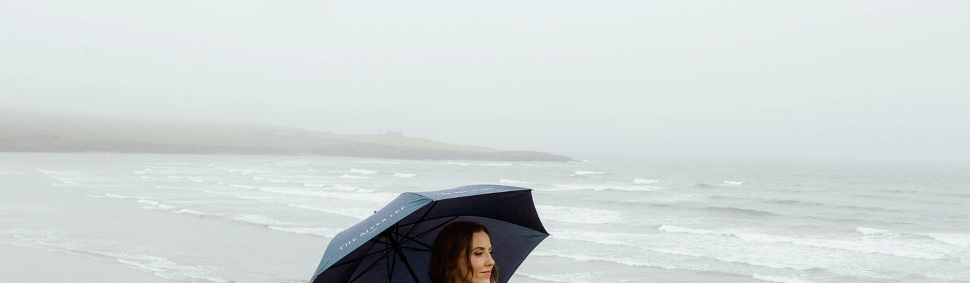 Louise O'Neill standing on the West Cork coast with an umbrella against a moody gray sky, showcasing the wild beauty of Cork's seaside.