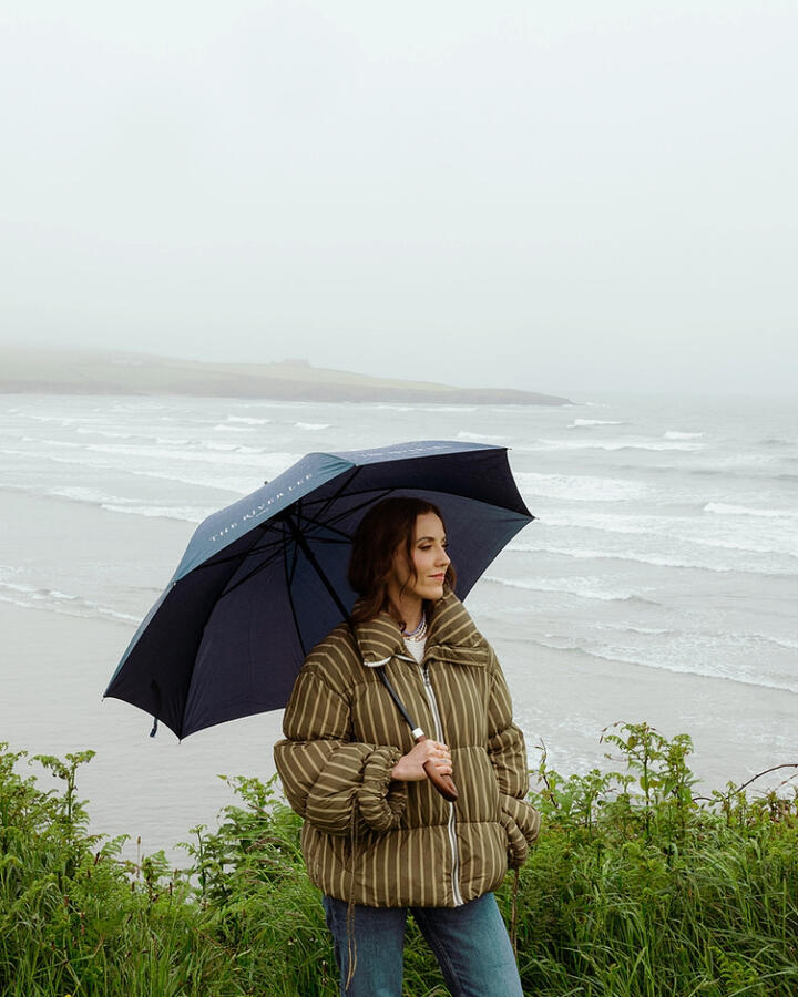 Louise O'Neill standing on the West Cork coast with an umbrella against a moody gray sky, showcasing the wild beauty of Cork's seaside.