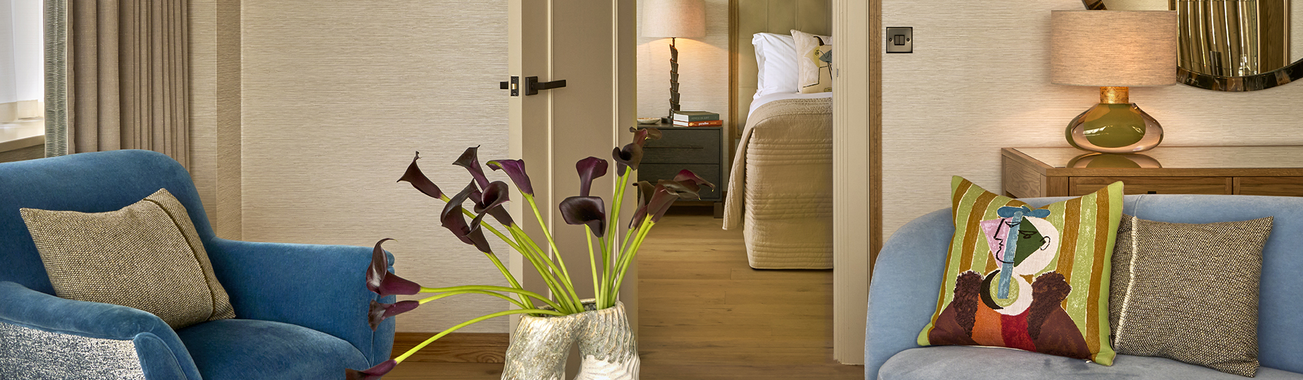 Living area of a suite at The River Lee hotel with a sofa, armchair, fresh flowers, and a king bed visible through an open door.